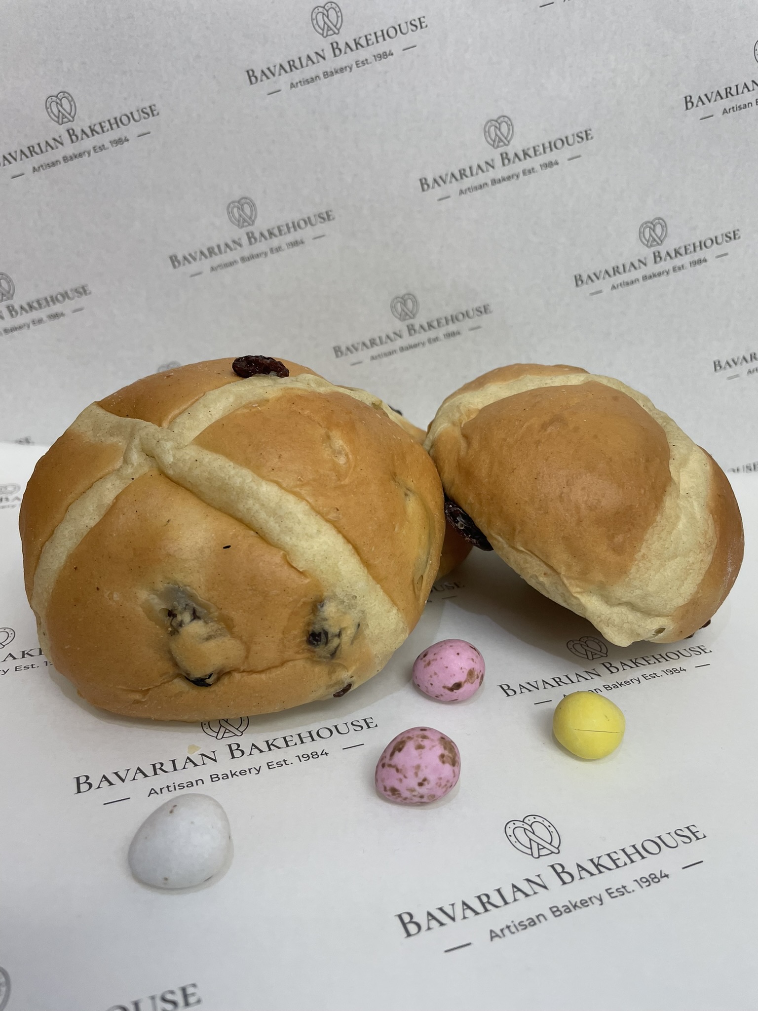 a spiced enriched dough bun filled with mixed peel and sultanas baked to a golden finish and topped with a white dough cross on top.