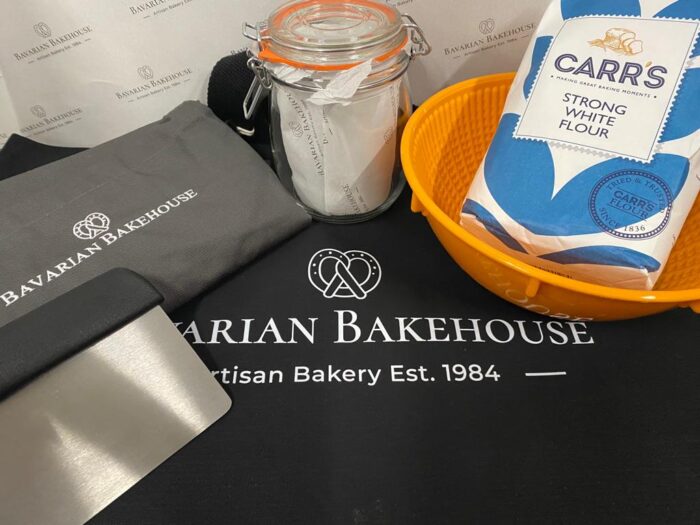 bakers goody bag: dark grey apron with logo, glass sourdough jar, plastic proving bowl, bag of carrs strong white flour, metal scraper