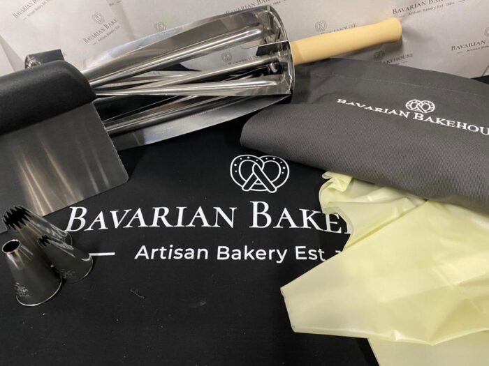 logo black apron, metal and wood croissant roller, reusable piping bag, 3 various piping nozzles, metal scraper and a large black cotton shopper bag with white bakery logo
