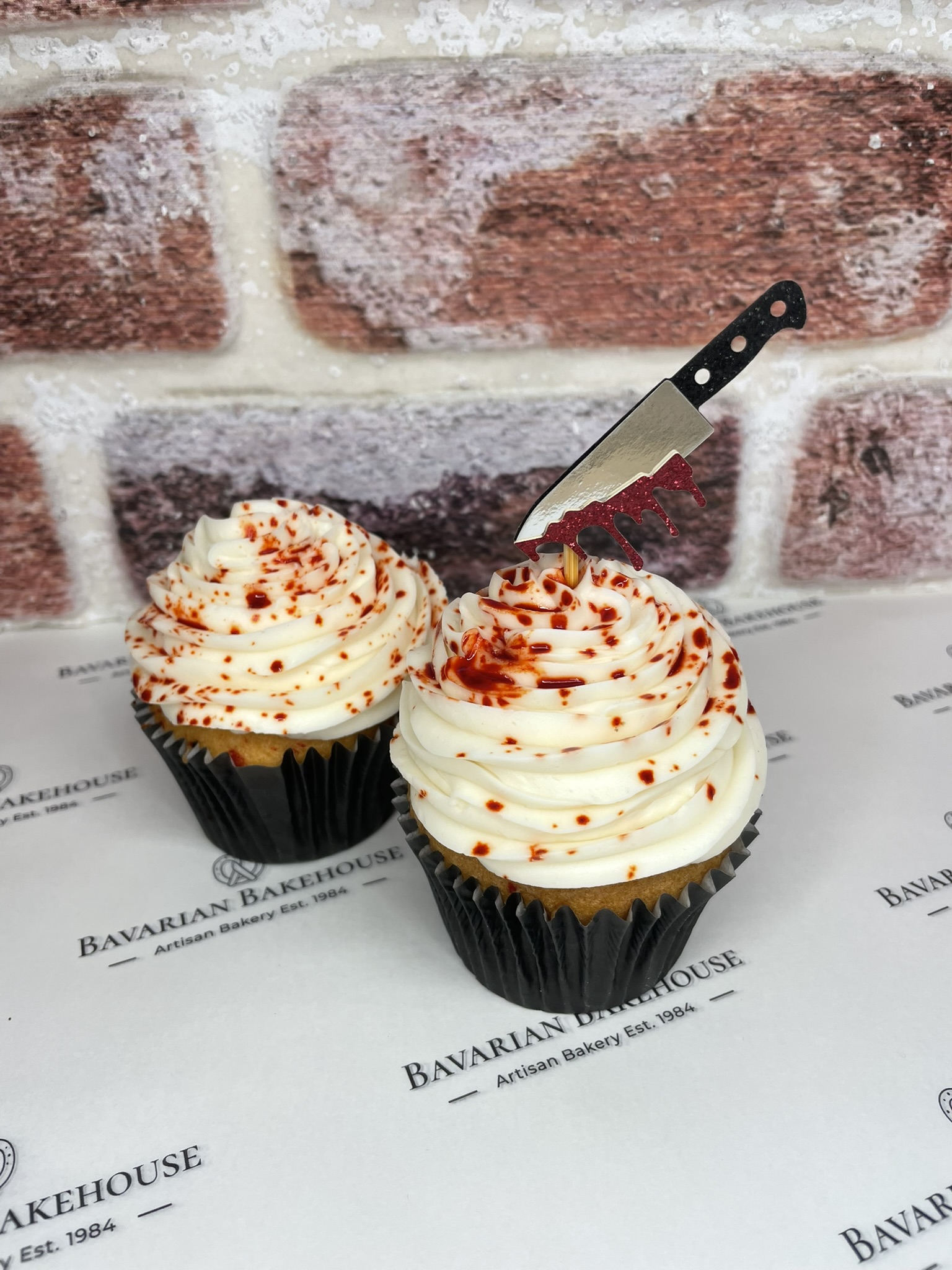 Vanilla cupcake with white frosting, decorated with red food colouring blood splatter and topped with a cut out paper knife.