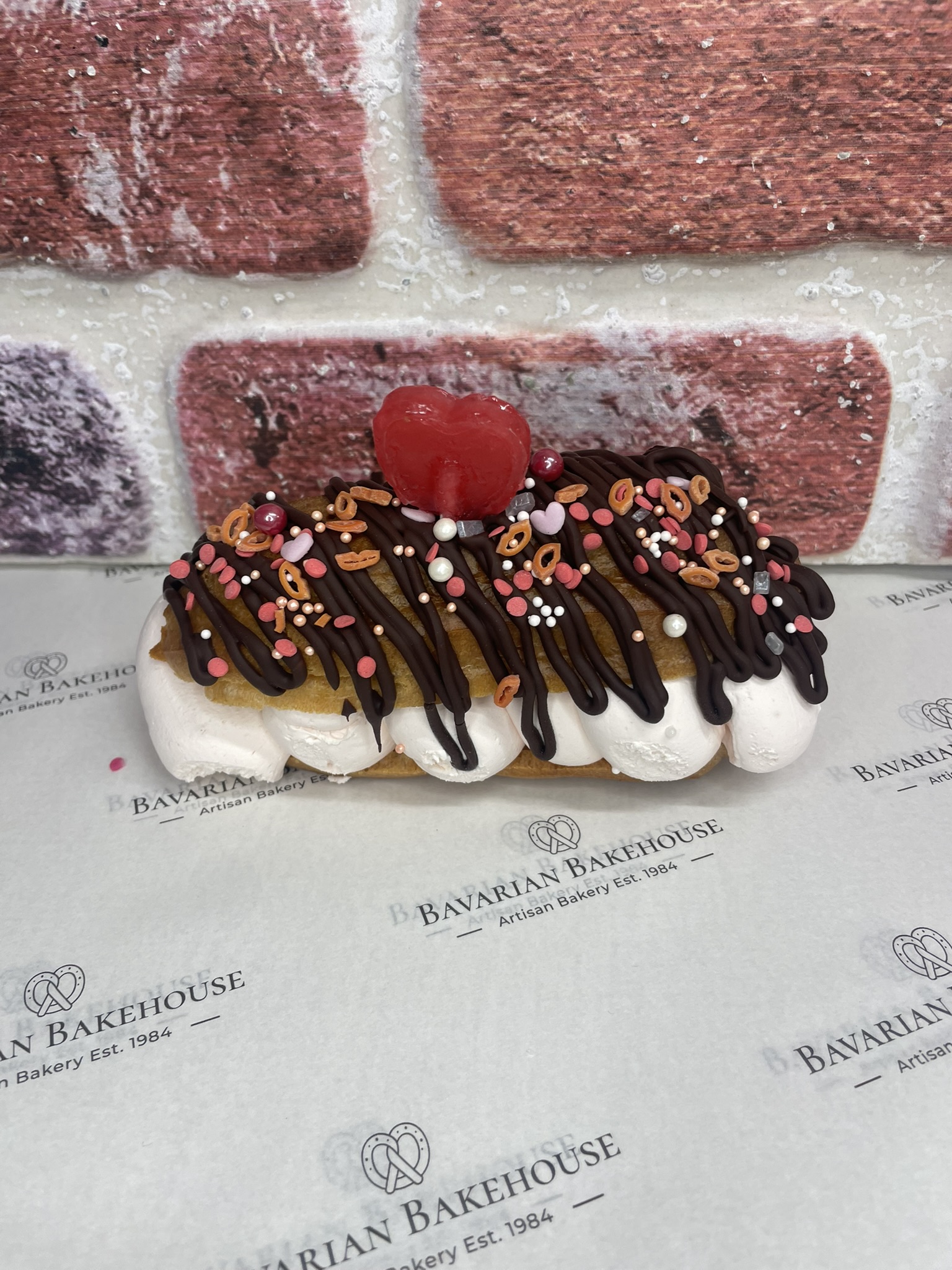 half sized artisan éclair filled with strawberry cream, topped with a dark chocolate drizzle, themed sprinkles and a heart shaped lolly.
