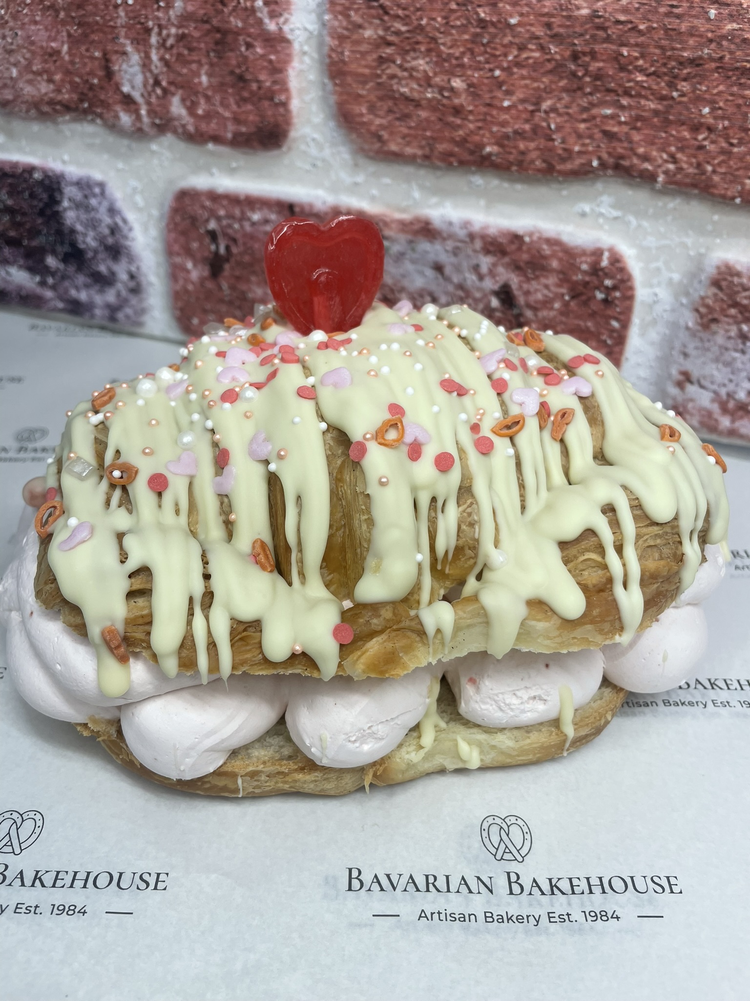 large handmade croissant filled with strawberry cream, topped with a shite chocolate drizzle, themed sprinkles and a hear lolly